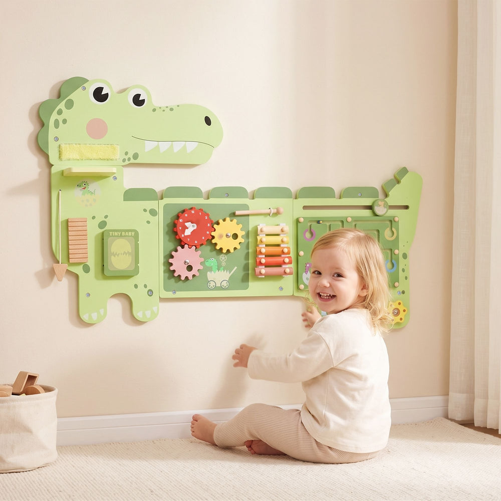 A child seated beside the green Qaba dinosaur busy board mounted on a playroom wall.